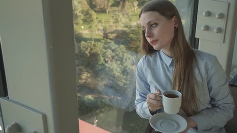 Concept: millennial, connections, lifestyle. Business casual dressed attractive girl drink tea by the window of trendy restaurant in skyscraper. HD 1080p. Slow motion 60fps