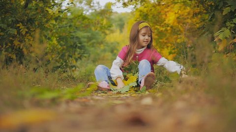 beautiful little girl with long hair collects yellow and green leaves sitting on the footpath. in the autumn in the city park. slow-motion