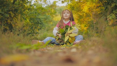 beautiful little girl with long hair tosses a yellow and green leaves up sitting on the footpath. Baby laughs. Autumn in the city park.