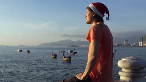 Man in the Christmas cap sits on the railing and looking at fishing boas in sea and raising sun. Asia. Vietnam. Christmas. New Year
