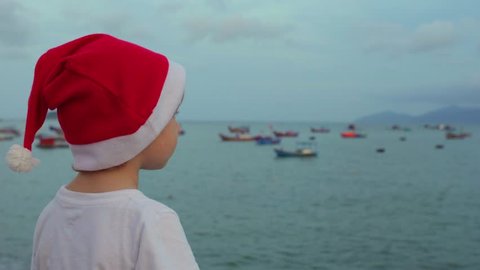 Cute boy in the Christmas cap sits on the railing and looking at fishing boas in sea. Asia. Vietnam. Christmas. New Year