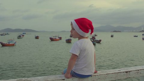 Cute boy in the Christmas cap sits on the railing and looking at fishing boas in sea. Asia. Vietnam. Christmas. New Year