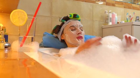Beautiful brunette woman wearing cosmetic face mask relaxes in foamy bath. Beauty treatment at home. 4K shot