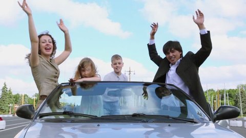 Parents and two kids stand in cabriolet and wave hands at sunny day