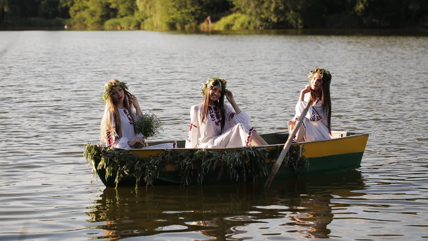 Three girls in the Slavic national costume in a boat floating on the river. Girls in wreaths posing and laughing merrily. Slavic girls in embroidered shirts.