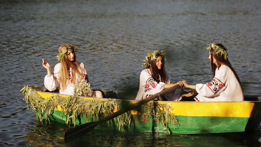 Three beautiful girls in the Slavic clothes in a boat floating on the river. Two girls rowing oars on the water. girl with long hair