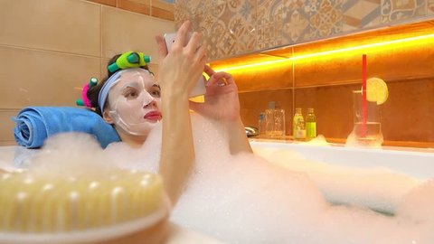 Brunette woman wearing face mask taps on her smartphone in foamy bathtub. 4K video