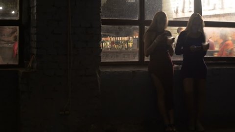 Two beautiful women drinking cocktails, browsing on smartphones at nightclub
