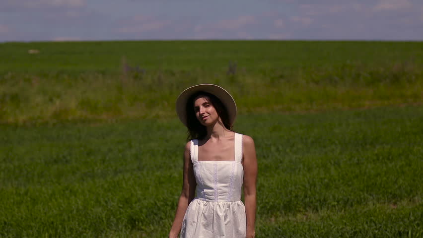 young girl in white dress and straw hat is on the green field with grass and smiles, holds the hand hat, summer. the model goes on camera