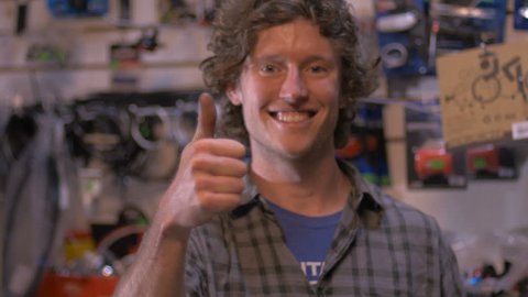 A handsome millennial small business owner smiles and gives a thumbs up in his retail bike shop