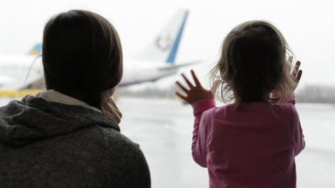 Close-up of attractive young mother and little daughter looking out the window at airport terminal, with airplane on the background.