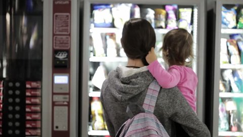 Young mother with little daughter selecting a snacks at vending machine inside airport