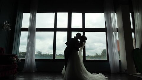 Confident groom meets happy bride first time seeing her in wedding dress silhouetted near window. Marrying couple embrace and kiss standing near huge window in room