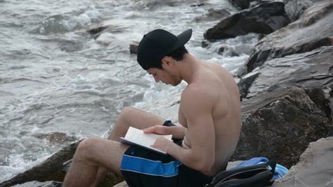 Shirtless Young Handsome Man Busy Reading a Book While Sitting on Beach Boulders next to Sea or Ocean
