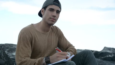 Shirtless Young Handsome Man Busy Writing on School Book While Sitting on Beach Boulders next to Sea or Ocean