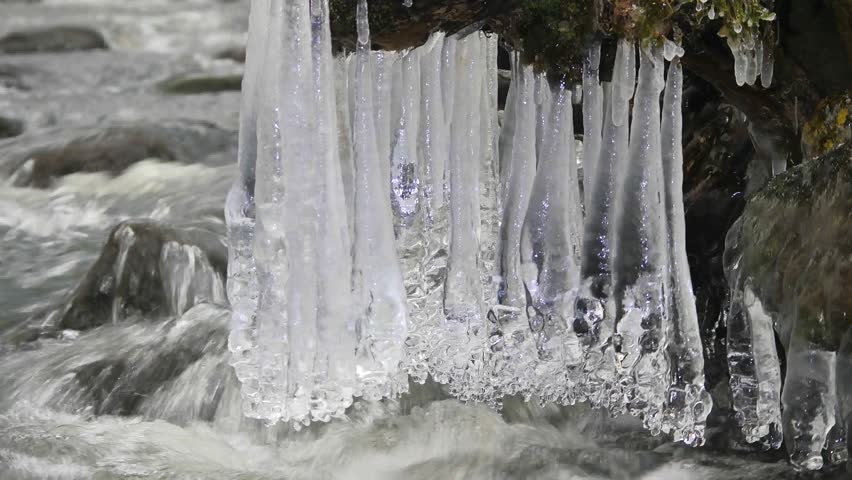 Long icicles hang above dark cold water of mountain river. Icicles glitter above milky water of stream. Branches of fallen trunks are covered by small flakes of powder snow. 4k 3840 X 2160 