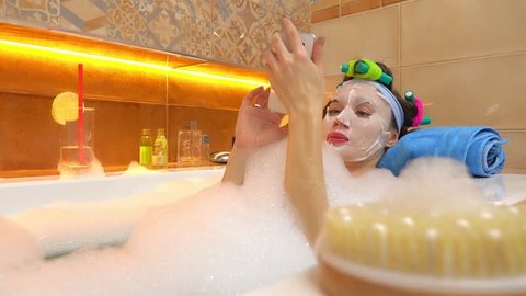 Brunette woman wearing face mask uses her mobile phone in foamy bathtub