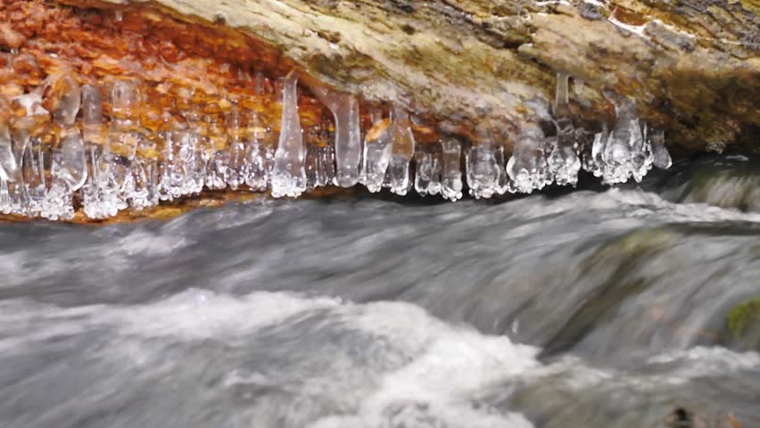 Shinning icicles. Frozen drops above winter stream created beautiful icicles.  Glittering ice above foamy winter brook. Fallen trunk and branch with ice cover and small snow flakes. 4k, 3840 X 2160