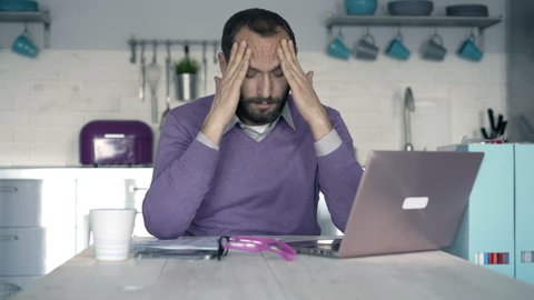 Young man having head pain while working on laptop by table in kitchen
