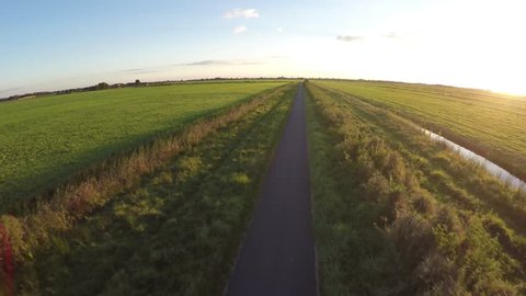 Aerial on beautiful summer evening with sunshine flying over straight small rural road moving fast forward green fresh grass colors on both sides of path and some bushes on right side with foliage 4k