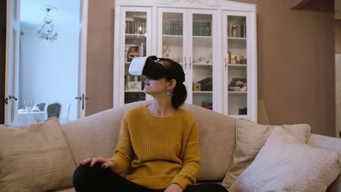 Beautiful young woman sitting on sofa at living room and using VR headset. 4K