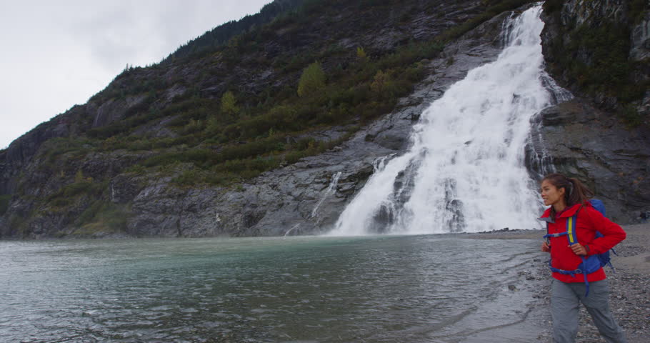 Alaska glacier landscape - tourist visiting Mendenhall Glacier tourist attraction. Woman hiking in front of famous cruise shore excursion near Juneau. Nugget Falls waterfall on left. RED EPIC SLOW MO.