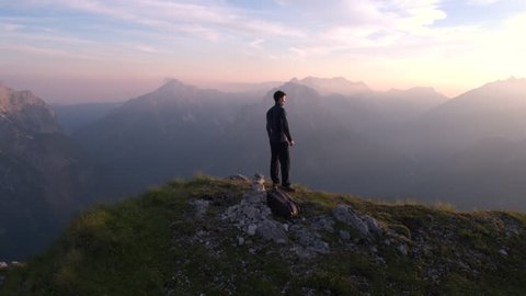 Aerial - Flying from front to behind the man standing on top of the mountain watching beautiful sunset over the peaks