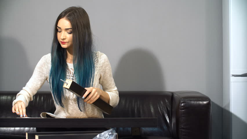 Young cute girl with colored hair sitting on a couch in their new apartment to put together a table. Shoot in kitchen. Following instructions is trying