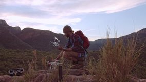 Male drone enthusiast, hiker preparing drone for arial flight in mountain range at sunset.