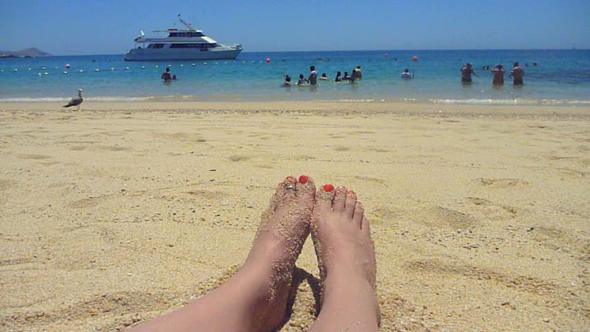 Woman's feet in sand with yacht in tropical location. Chileno Bay in Cabo,