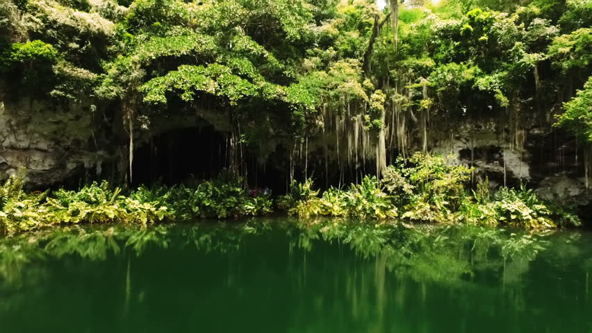 The camera flies to the entrance of the cave above the lake. Around the green trees