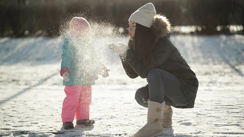 Young Mother is Blowing on the Snow in Her Hands, Little Daughter is Looking on Snowflakes. Woman with Baby Girl Playing Outdoors in the Morning in Cold Sunny Weather at Winter Time.