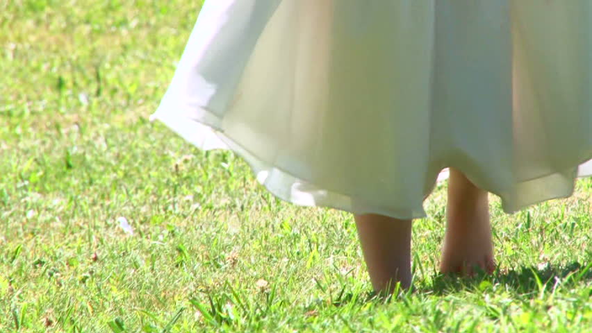 Young little girl in white dress walks bare footed on green grass in summer.