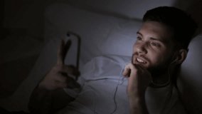technology, internet, communication and people concept - happy smiling young man with smartphone and earphones having video conference and talking to someone in bed at night