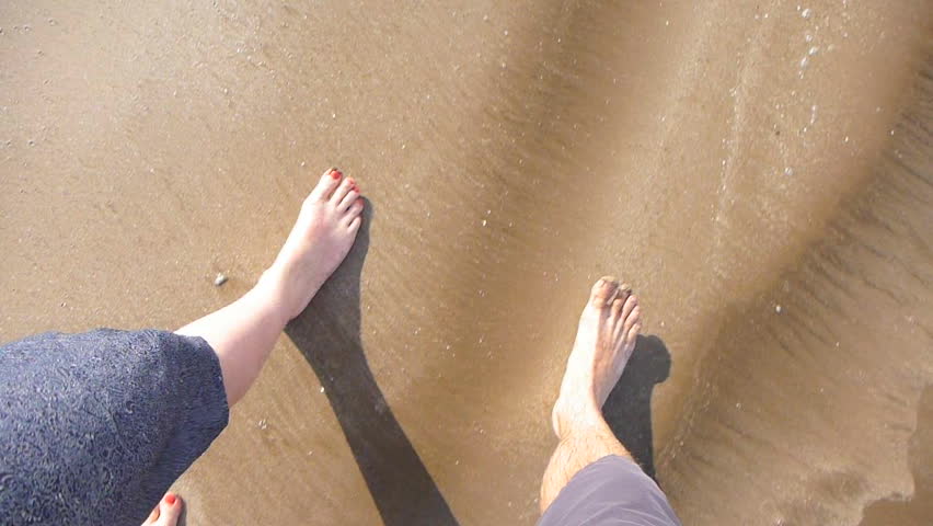 Man and woman's feet point of view while walking on sandy beach with waves