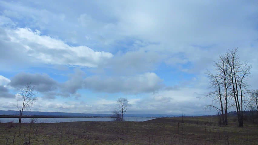 Time lapse of party cloudy skies over grassland with bare trees near river in