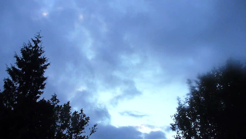 Clouds move fast, toward camera at dusk with silhouetted trees.