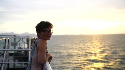 beautiful young girl with short hair wearing glasses and overalls posing at the camera standing on the deck of the ship. sea sunset background.