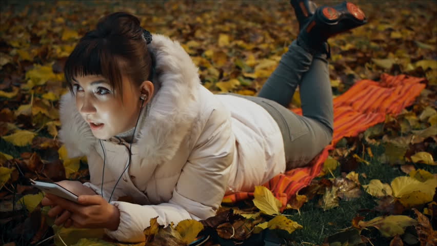 Young woman intently talking on the phone through headphones lying on fallen leaves in autumn park.