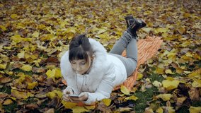 Pretty woman with attractive makeup starts video call lying on yellow and red fallen leaves in autumn park. Face lights up with smartphone screen light. Midshot.