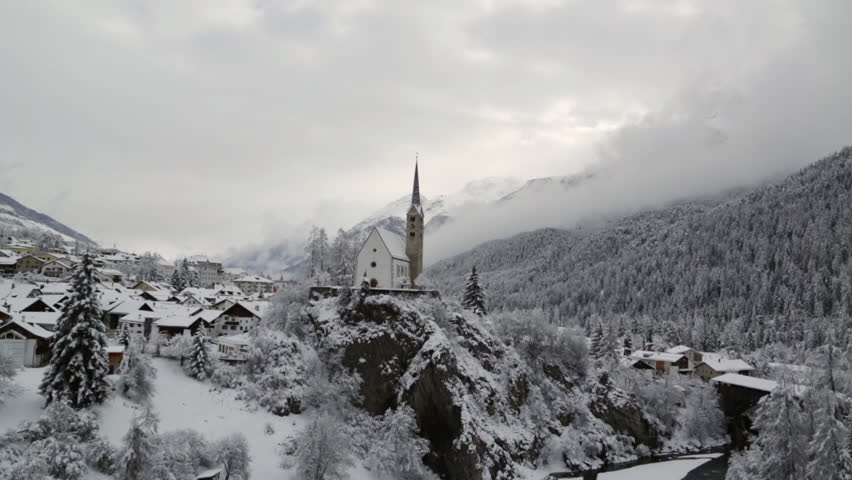 A video overlooking the reformed church and the Inn River in Scuol, Graubünden, Switzerland. The river is flowing beneath the church against a peaceful winter landscape. 