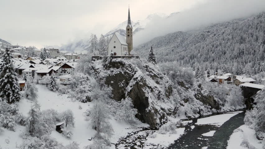 A video overlooking the reformed church and the Inn River in Scuol, Graubünden, Switzerland. The river is flowing beneath the church against a peaceful winter landscape. 