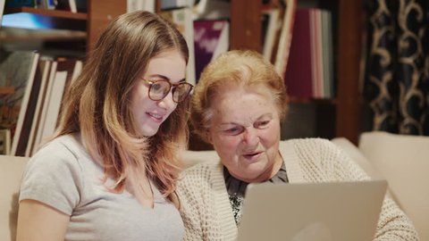 Beautiful old woman and young girl are using a laptop, talking and smiling while sitting on couch at home
