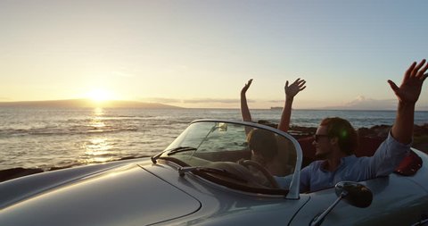 Happy Couple Driving on Country Road By the Ocean into the Sunset in Classic Vintage Sports Car. Hands in the Air. Shot on RED