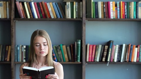 Cute teen girl learn close up. Smart 20s blonde model read a literature turning pages of the book holding hands closeup. Shelves with books on background and inspiration. Natural lighting indoors