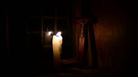 A candle burns on the window. Outside night. The cross with a crucifix standing behind. Candle flame flickers and dies. Wooden windows frame in a wooden house in the mountains. 