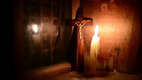 A candle burns on the window. Outside night. The cross with a crucifix standing behind. (Focus on the cross). Candle flame flickers. Wooden windows frame in a wooden house in the mountains. 