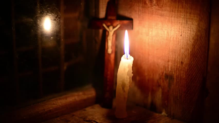 A candle burns on the window. Outside night. The cross with a crucifix standing behind. (Focus on candle). Candle flame flickers. Wooden windows frame in a wooden house in the mountains. 