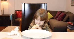 4k video. Three years old blonde child with green shirt eating pizza piece with her hands, cleaning with napkin, sitting in dark brown chair in table with grey tablecloth at home
