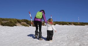 4k video. Three years old blonde child and woman mother with white and purple coats walking on snow in winter mountain from backwards together

